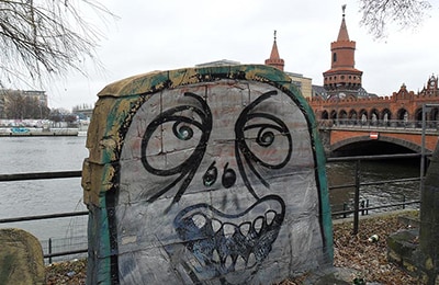 Reste einer Mauer, auf die ein karikaturhaftes Gesicht gesprayed ist. Im Hintergrund die Spree.