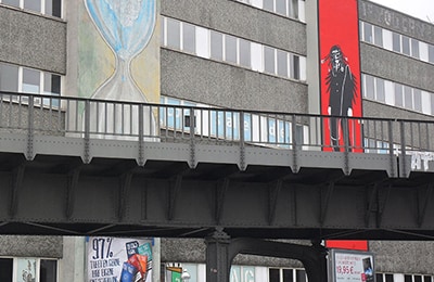 Ein graues Gebäude, an dem zwei große Kunstwerke befestigt sind. Das linke zeigt eine Sanduhr und das rechte eine maskierte Gestalt vor roten Hintergrund.