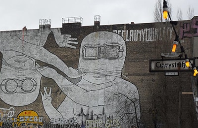 Riesiges Graffiti an Hauswand, das zwei verkleidete Gestalten zeigt, die sich gegenseitig die Maske abziehen.