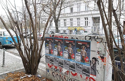 Stromkasten am Straßenrand mit Plakaten vollgeklebt, im Hintergrund ein weißes Reihenhaus.