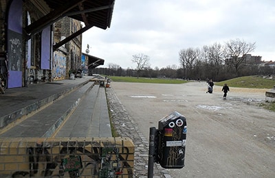 Front einer altenbesprayten Lagerhalle, die an ein offenes Gelände grenzt.