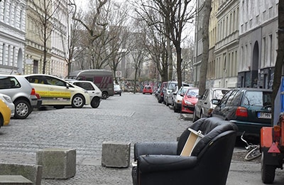 Straße in Kreuzkölln, auf deren beiden Seiten Autos parken. Im Vordergrund steht ein schwarzer Sessel und weiterer Sperrmüll.